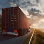 Semi-truck with cargo container driving on a coastal road at sunset.