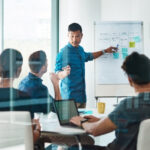 Business team in a meeting room discussing strategy and crisis planning, with a presenter pointing to notes on a whiteboard.