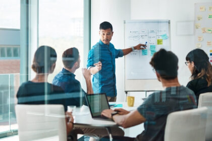 Business team in a meeting room discussing strategy and crisis planning, with a presenter pointing to notes on a whiteboard.