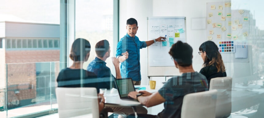 Business team in a meeting room discussing strategy and crisis planning, with a presenter pointing to notes on a whiteboard.