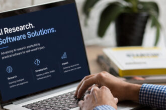 Laptop screen showing AI research and software solutions messaging, with a person working beside a coffee cup and notebook.