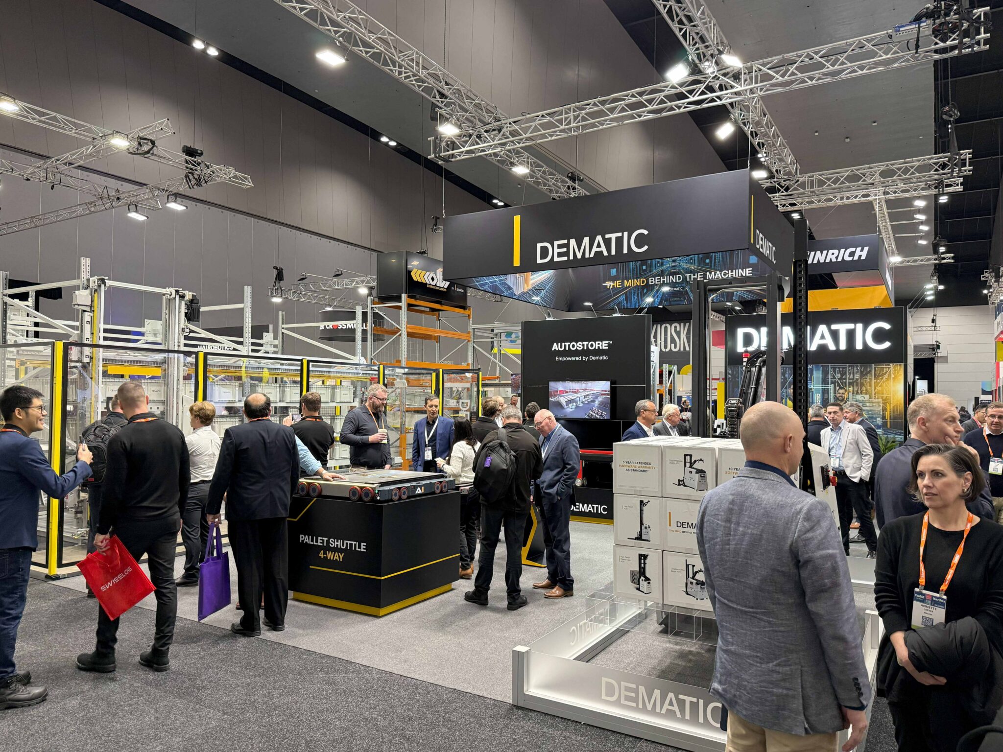 Visitors gather at the Dematic stand during CeMAT Australia 2026, where live demonstrations of warehouse automation technologies, including AutoStore and a 4-way pallet shuttle system, attract attendees exploring intelligent supply chain solutions. Large Dematic branding and exhibition displays are visible throughout the busy trade show floor.