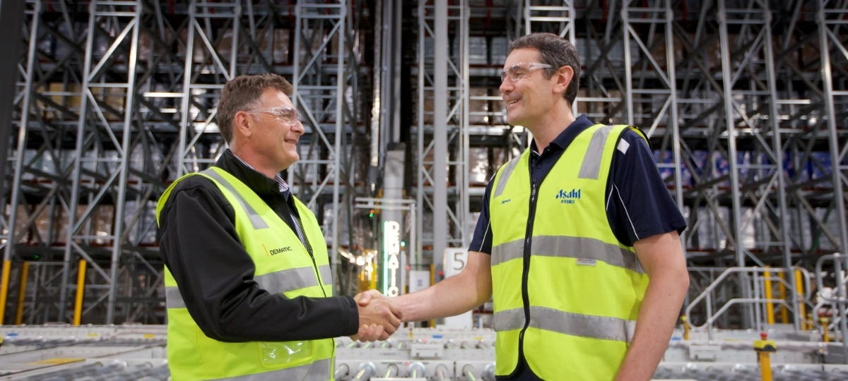 Dematic and Asahi Beverages representatives shaking hands inside an automated warehouse facility, marking a partnership milestone.