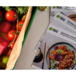 HelloFresh meal kit box filled with fresh produce, including tomatoes and herbs, alongside recipe cards featuring a selection of prepared meal options.