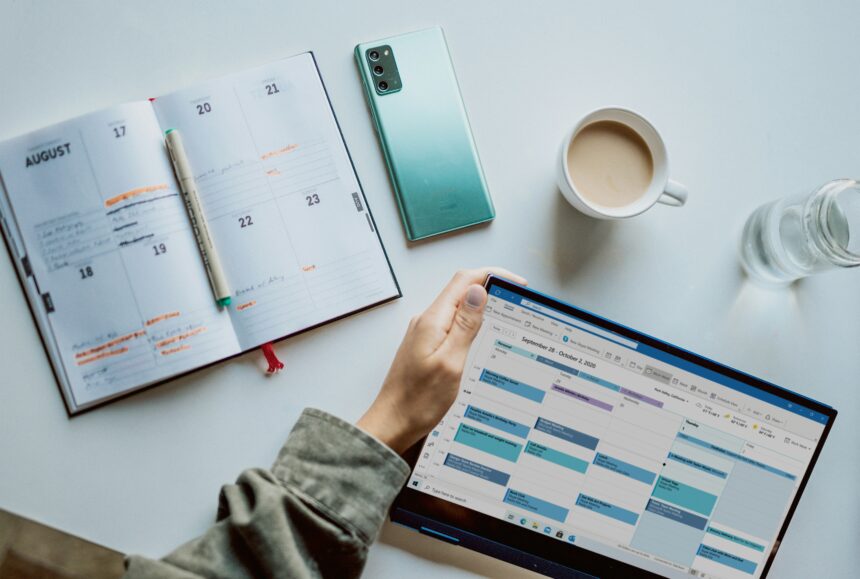 Overhead view of a tidy workspace featuring an open weekly planner with handwritten notes, a pen, a smartphone, a tablet displaying a digital calendar, a cup of coffee and a glass of water, illustrating the use of both paper and digital tools for scheduling and time management.