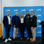 Five people stand together in front of a blue JAS-branded media wall at an event, flanked by blue and white balloon displays, with a lectern positioned to the side.