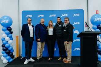 Five people stand together in front of a blue JAS-branded media wall at an event, flanked by blue and white balloon displays, with a lectern positioned to the side.