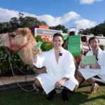 Two men in white bathrobes pose beside a camel carrying green Two Dudes products outside a Coles store, celebrating a promotional delivery with product boxes loaded on the camel.
