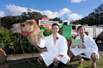 Two men in white bathrobes pose beside a camel carrying green Two Dudes products outside a Coles store, celebrating a promotional delivery with product boxes loaded on the camel.