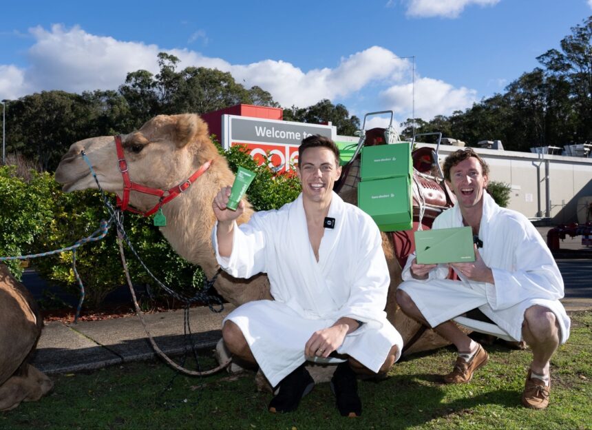 Two men in white bathrobes pose beside a camel carrying green Two Dudes products outside a Coles store, celebrating a promotional delivery with product boxes loaded on the camel.