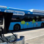 A blue zero-emissions bus is parked beside a Zenobē electric charging unit, with charging cables connected in a depot setting.