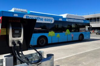 A blue zero-emissions bus is parked beside a Zenobē electric charging unit, with charging cables connected in a depot setting.