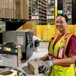 A warehouse worker wearing a high-visibility safety vest, gloves and safety glasses places a sealed cardboard package at a packing station beside a label printer. High-bay racking, stacked cartons and pallets are visible in the background of the distribution centre.