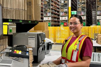 A warehouse worker wearing a high-visibility safety vest, gloves and safety glasses places a sealed cardboard package at a packing station beside a label printer. High-bay racking, stacked cartons and pallets are visible in the background of the distribution centre.