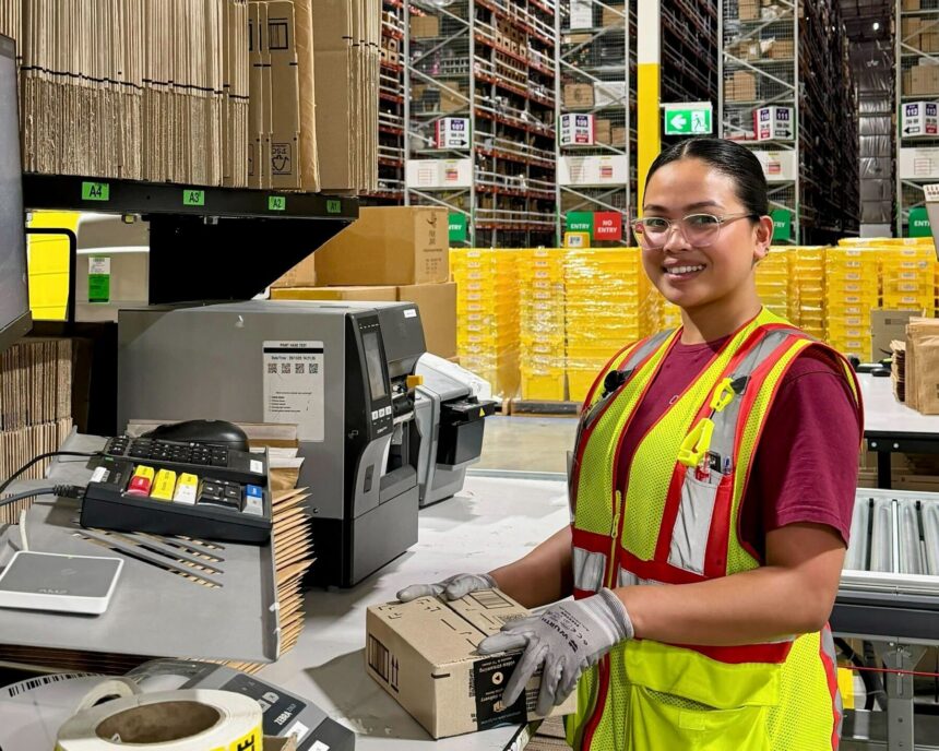A warehouse worker wearing a high-visibility safety vest, gloves and safety glasses places a sealed cardboard package at a packing station beside a label printer. High-bay racking, stacked cartons and pallets are visible in the background of the distribution centre.