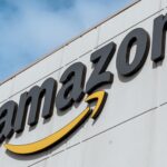 Close-up of the Amazon logo mounted on the exterior of a warehouse or fulfilment centre building against a blue sky.