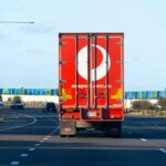 Rear view of an Australia Post delivery truck travelling along a multi-lane road, with the company's red branding visible on the trailer and light traffic ahead under a clear blue sky.