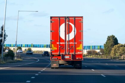 Rear view of an Australia Post delivery truck travelling along a multi-lane road, with the company's red branding visible on the trailer and light traffic ahead under a clear blue sky.