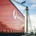 An Australia Post semi-trailer truck travels across a cable-stayed bridge alongside traffic, with Australian flags flying overhead under a partly cloudy sky.