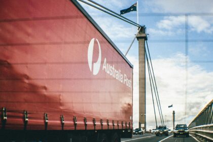 An Australia Post semi-trailer truck travels across a cable-stayed bridge alongside traffic, with Australian flags flying overhead under a partly cloudy sky.