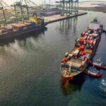 Aerial view of a busy container port with cargo ships, stacked shipping containers and towering gantry cranes, as tugboats guide a container vessel into position at the terminal.