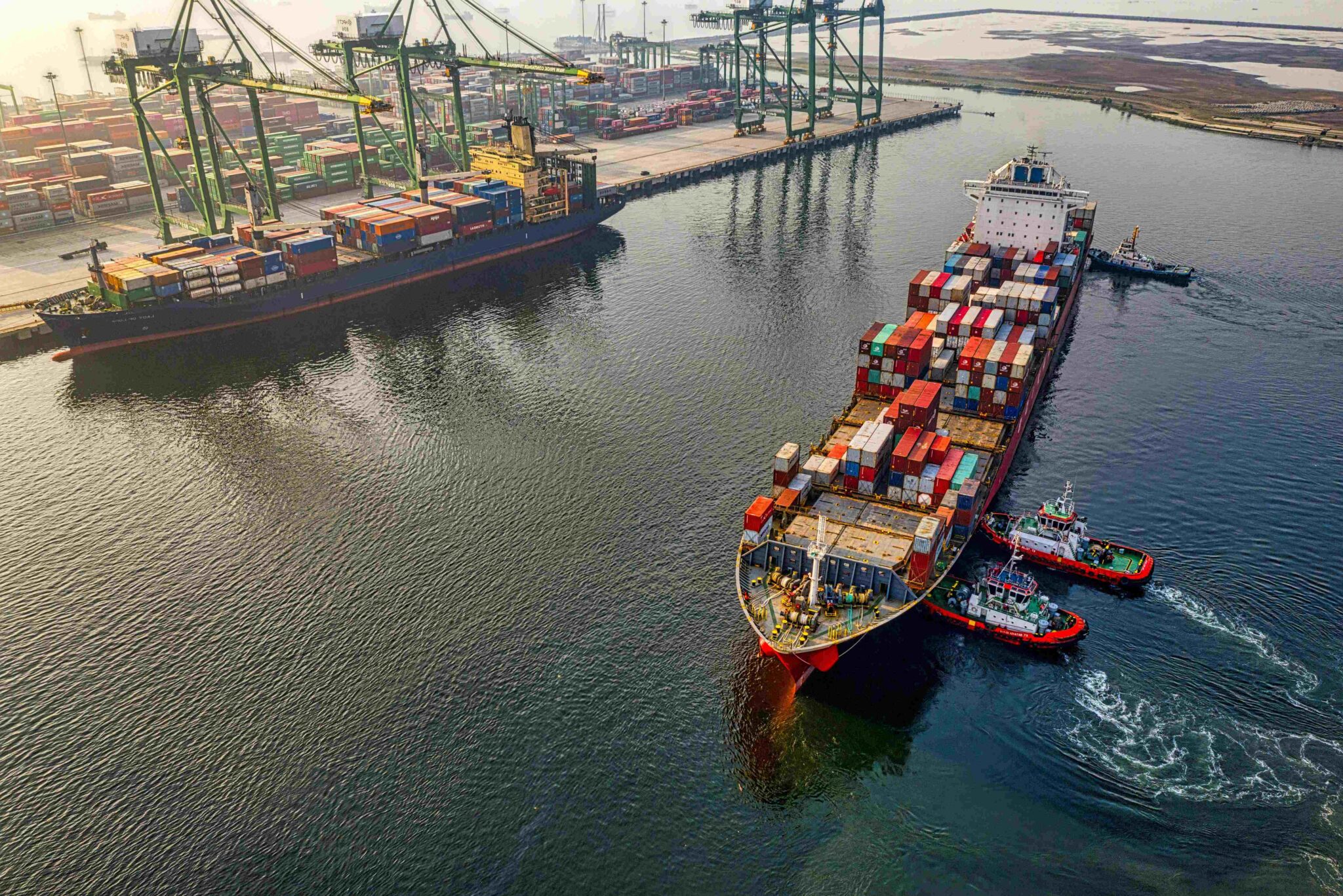 Aerial view of a busy container port with cargo ships, stacked shipping containers and towering gantry cranes, as tugboats guide a container vessel into position at the terminal.