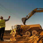 Caucasian Male Real Estate Investor And Civil Engineer Talking On Construction Site Of Apartment Block. Colleagues Discussing Building Progress. Excavator Loading Sand In Industrial Truck On Warm Day.