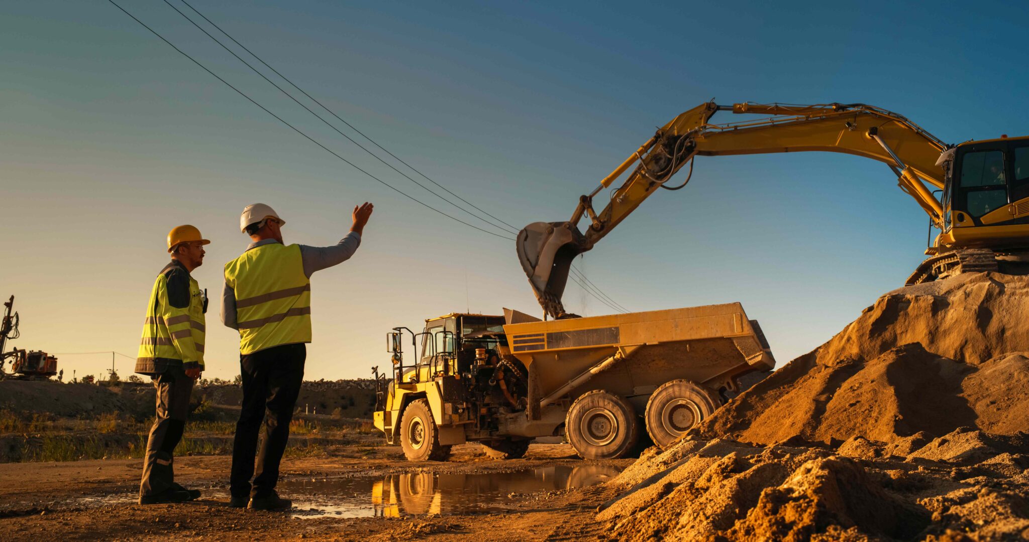 Caucasian Male Real Estate Investor And Civil Engineer Talking On Construction Site Of Apartment Block. Colleagues Discussing Building Progress. Excavator Loading Sand In Industrial Truck On Warm Day.