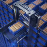 An automated shuttle system retrieves a blue storage tote from a high-density warehouse storage grid filled with stacked containers, illustrating automated goods-to-person fulfilment technology.
