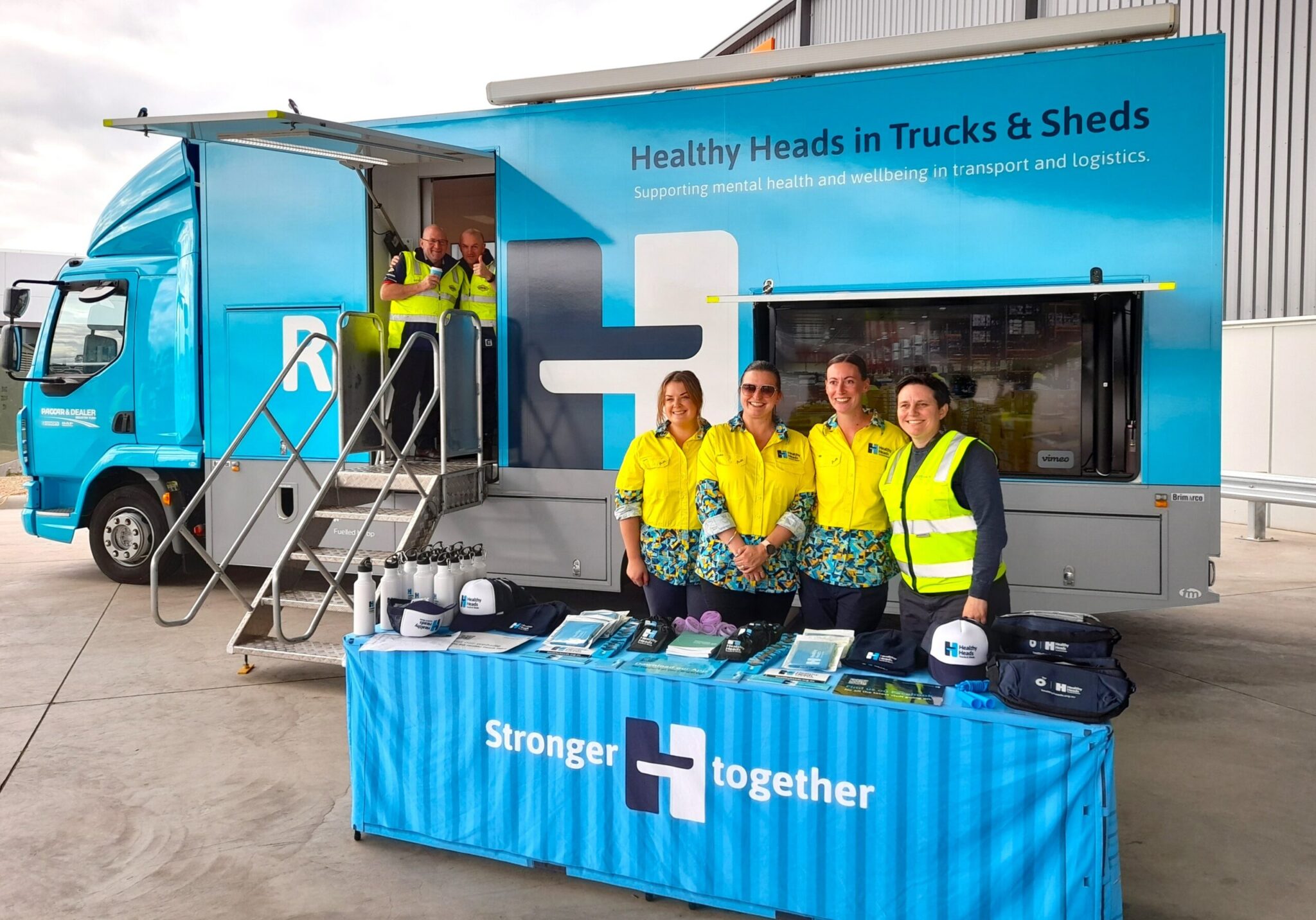 Healthy Heads in Trucks & Sheds mobile wellbeing truck with staff standing behind an information table displaying mental health resources for the transport and logistics industry.