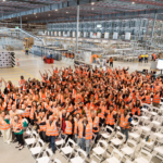 Hundreds of warehouse employees wearing high-visibility safety vests gather inside a large automated distribution centre, standing in front of conveyor systems, robotics and high-density storage infrastructure during a team event.