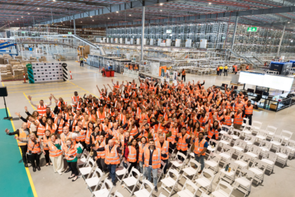Hundreds of warehouse employees wearing high-visibility safety vests gather inside a large automated distribution centre, standing in front of conveyor systems, robotics and high-density storage infrastructure during a team event.