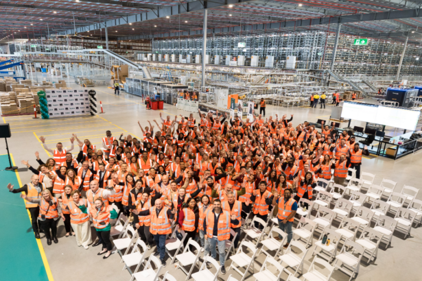 Hundreds of warehouse employees wearing high-visibility safety vests gather inside a large automated distribution centre, standing in front of conveyor systems, robotics and high-density storage infrastructure during a team event.