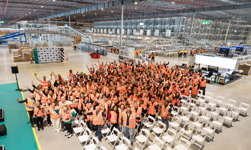 Hundreds of warehouse employees wearing high-visibility safety vests gather inside a large automated distribution centre, standing in front of conveyor systems, robotics and high-density storage infrastructure during a team event.
