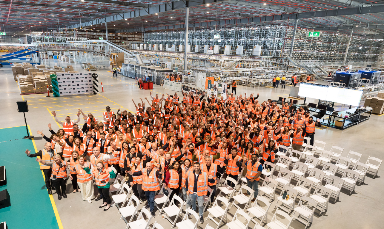 Hundreds of warehouse employees wearing high-visibility safety vests gather inside a large automated distribution centre, standing in front of conveyor systems, robotics and high-density storage infrastructure during a team event.