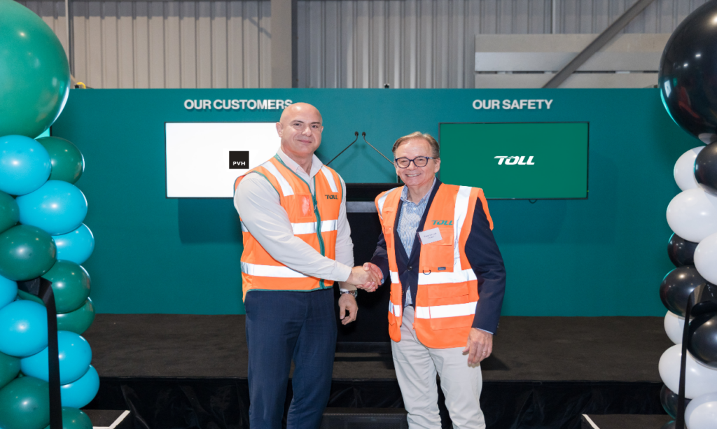 Two people wearing high-visibility safety vests shake hands on a stage inside a warehouse facility, with Toll branding, customer and safety displays, and balloon decorations in the background.