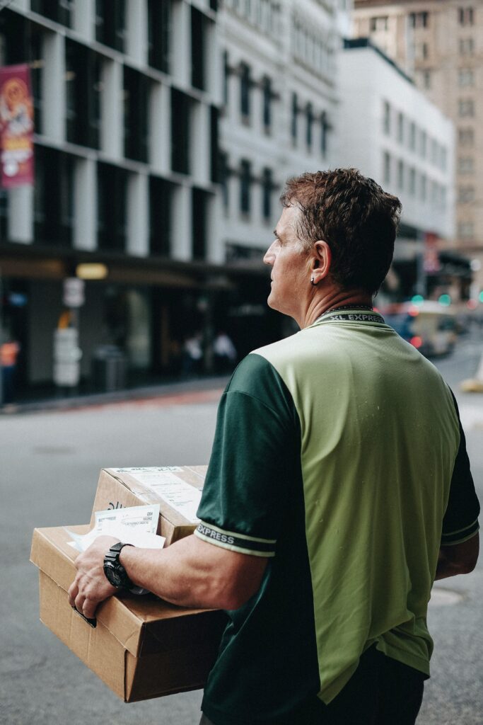 A delivery driver carries cardboard parcels while walking through a city street, preparing to complete a last-mile delivery in an urban business district.