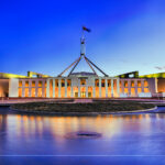 Australia's Parliament House in Canberra illuminated at dusk, with lights reflecting across the foreground water beneath a dark, cloud-filled evening sky.