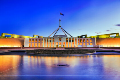 Australia's Parliament House in Canberra illuminated at dusk, with lights reflecting across the foreground water beneath a dark, cloud-filled evening sky.