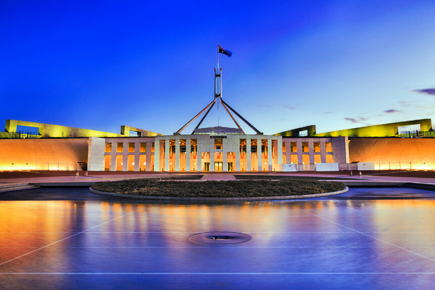 Australia's Parliament House in Canberra illuminated at dusk, with lights reflecting across the foreground water beneath a dark, cloud-filled evening sky.