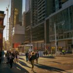 Pedestrians cross a busy Melbourne CBD intersection during the late afternoon, surrounded by high-rise office buildings, tram lines and city traffic in golden sunlight.