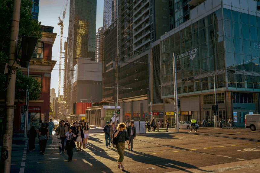Pedestrians cross a busy Melbourne CBD intersection during the late afternoon, surrounded by high-rise office buildings, tram lines and city traffic in golden sunlight.