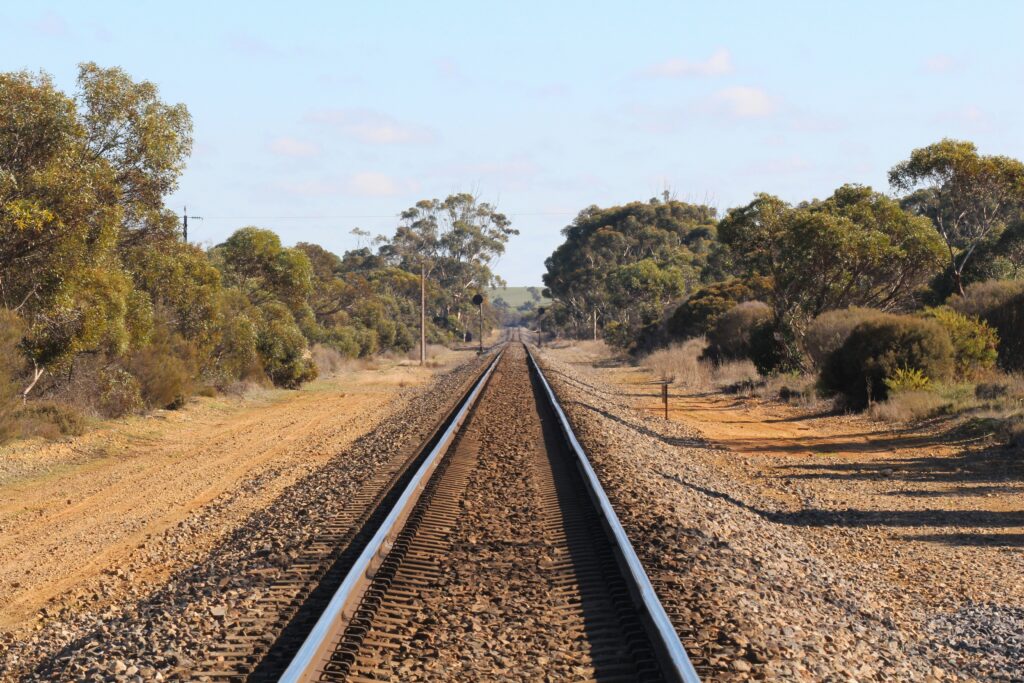 Railway tracks stretch into the distance through the Australian countryside, bordered by gravel, dirt access tracks and native bushland under a clear blue sky.