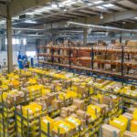 Wide view of a warehouse filled with shelving, yellow storage bins and cardboard boxes, with pallet racking and inventory storage extending across the facility.