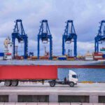 A container truck drives along a port terminal in front of a cargo ship and towering gantry cranes, illustrating the connection between road freight and international shipping.
