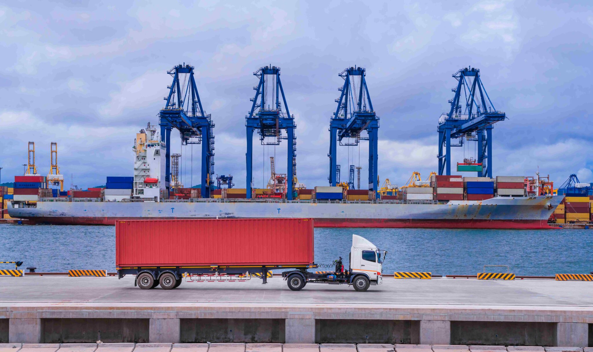 A container truck drives along a port terminal in front of a cargo ship and towering gantry cranes, illustrating the connection between road freight and international shipping.