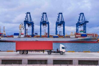 A container truck drives along a port terminal in front of a cargo ship and towering gantry cranes, illustrating the connection between road freight and international shipping.