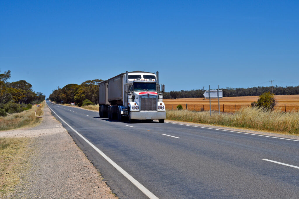 A semi-trailer truck travels along a two-lane highway through rural Australia, with open farmland, native trees and a clear blue sky stretching into the distance.