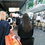 Two shoppers walk along a busy city shopping street carrying retail bags, with cafés, pedestrians and public transport visible in the background.