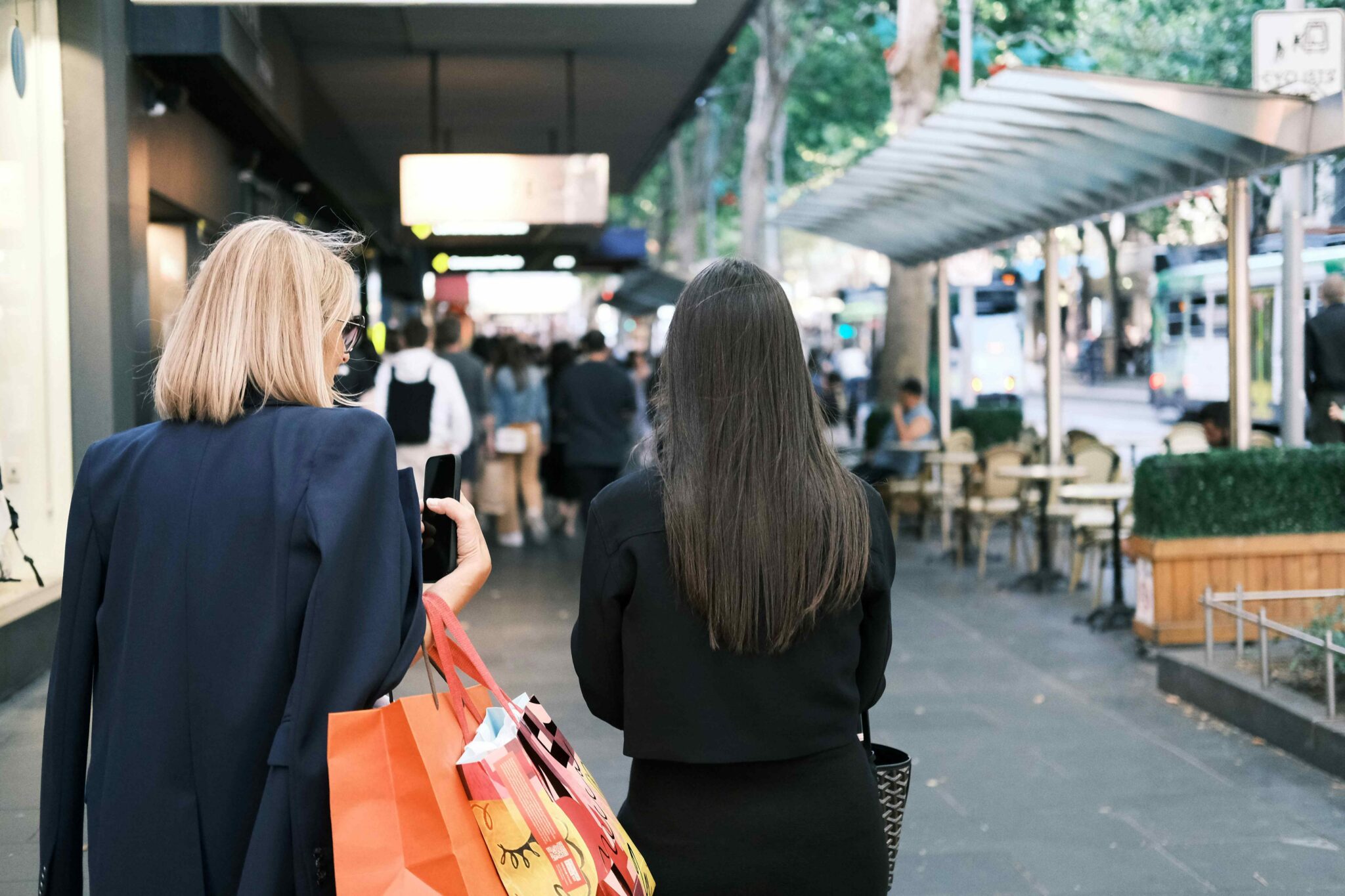 Two shoppers walk along a busy city shopping street carrying retail bags, with cafés, pedestrians and public transport visible in the background.